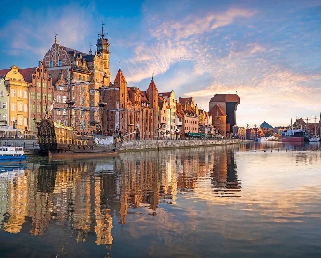 Gdańsk’s Motława waterfront at sunset, with colourful merchant houses, a tall ship and the medieval crane reflected in the river